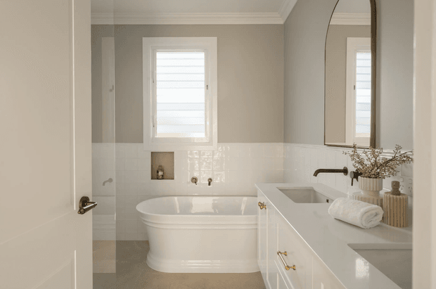 A bright, refined bathroom viewed through an open white door. The room features a freestanding white bathtub beneath a shuttered window, white glossy wall tiles, a pale grey painted upper wall, and a white vanity with integrated basins, brass hardware, and a dark metal wall-mounted tap. A large arched mirror and neatly styled towels and accessories add a classic yet contemporary character.