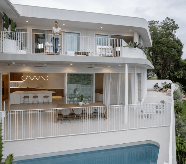 A bright, multi-level contemporary residence viewed from the exterior, featuring rounded white balcony slabs, white vertical balustrades, extensive glazing, and a swimming pool in the foreground. The lower level opens to a covered outdoor dining and entertaining area beside a warm-toned kitchen with an island and stools, while the upper balcony includes outdoor seating. Lush landscaping surrounds the building.