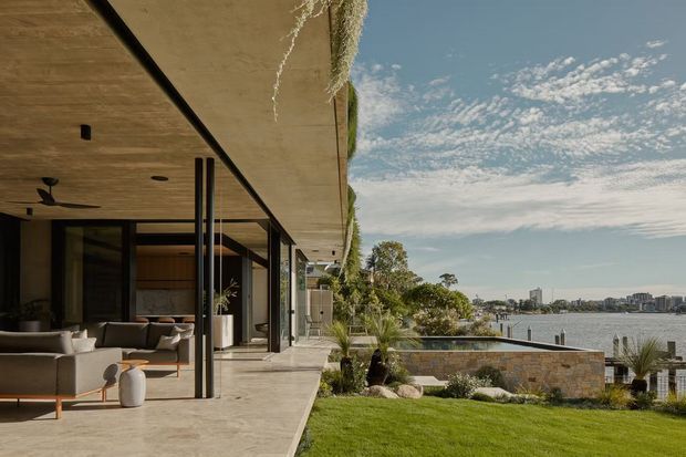 A contemporary waterfront residence viewed from the landscaped garden, with a broad concrete cantilevered roof and terrace extending over full-height glazed living spaces. The scene includes an outdoor pool or water feature, stone retaining wall, lawn, planted garden, and a calm urban harbour setting beyond.
