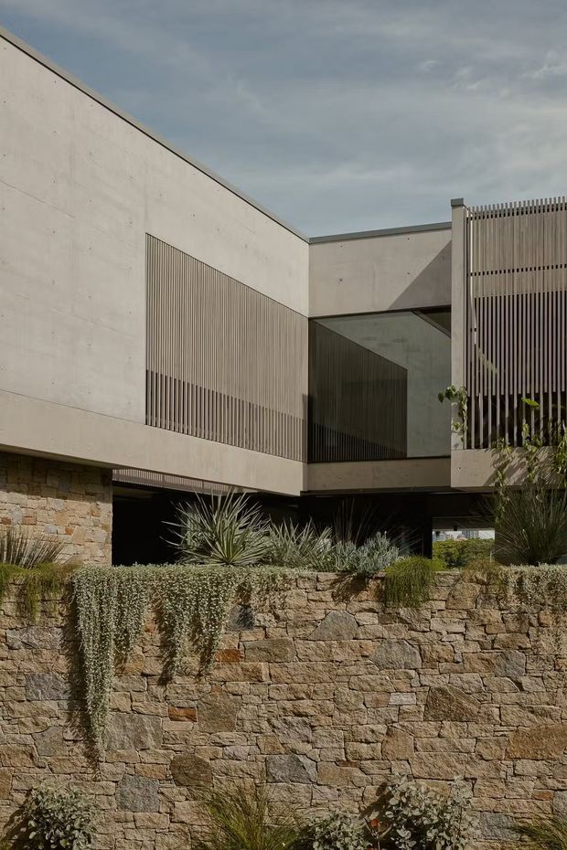 A contemporary two-storey architectural exterior with a pale concrete-like rendered facade, strong angular roofline, recessed dark glazing, and vertical metal screening. A substantial stacked-stone retaining wall and drought-tolerant landscaping sit in the foreground, creating a modern, textural residential composition.