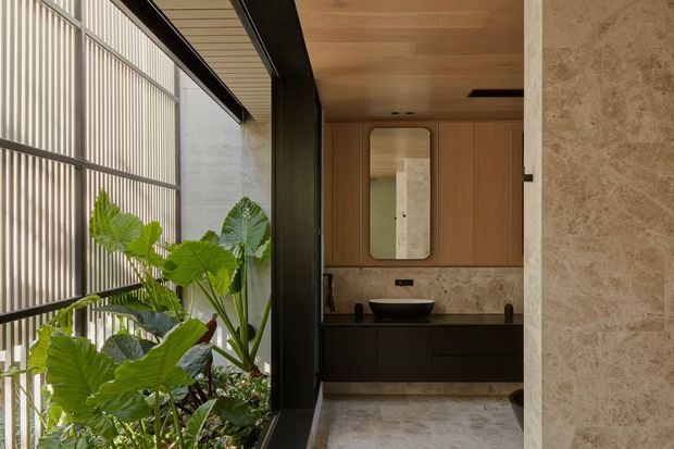 A refined contemporary bathroom viewed through a glazed opening, featuring a dark floating vanity with vessel basin, timber wall cabinetry, a rounded-rectangle mirror, marble or stone wall and floor finishes, and a full-height window with vertical screening beside lush indoor planting. The space has a warm, natural material palette and strong connection to greenery and daylight.