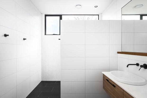 Bright contemporary bathroom with large white wall tiles, dark charcoal floor tiles, a walk-in shower area, wall-mounted matte-black tapware, and a timber-look floating vanity with a white basin. A large mirror and high horizontal window add spaciousness and natural light.