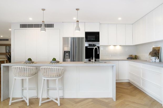 Bright, spacious white contemporary kitchen with a large paneled island, two white bar stools, integrated cabinetry, stainless-steel refrigerator, built-in black appliances, pendant lights, and a light herringbone timber floor. The kitchen has a clean, refined appearance with ample storage and subtle warm accents.