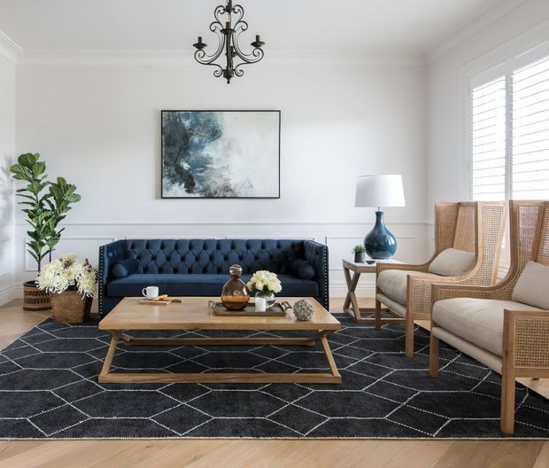 A bright, refined living room with white paneled walls, decorative cornices, plantation shutters, a dark tufted sofa, woven cane armchairs, a timber coffee table, patterned dark rug, and a traditional chandelier. Blue and neutral furnishings are balanced with greenery and restrained artwork.