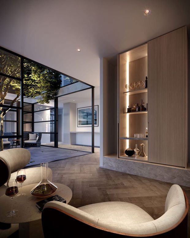 Sophisticated contemporary living room with a sculptural upholstered lounge chair and round coffee table in the foreground. A built-in timber bar cabinet with illuminated open shelving and concealed doors forms a feature wall, while black-framed glass partitions open onto an internal courtyard with planting and additional seating. Warm layered lighting, neutral finishes, and a herringbone timber floor create an elegant, inviting atmosphere.