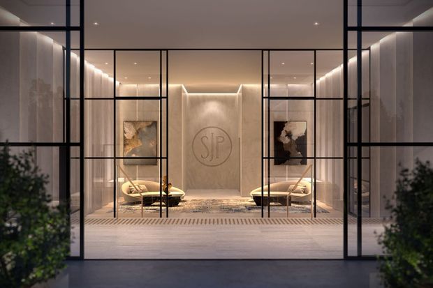 A refined, symmetrical entrance lobby or reception space viewed through large black-framed glazed doors. The interior features warm concealed and recessed lighting, pale textured wall finishes, matching lounge chairs, abstract artwork, a central circular monogram feature, and a patterned rug. The dark metal grid framing contrasts with the soft neutral interior, creating a polished contemporary hospitality or commercial atmosphere.