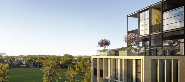 Exterior architectural view of a contemporary multi-storey building overlooking a broad green park or landscaped open space. The facade combines extensive floor-to-ceiling glazing, dark metal framing, projecting balconies or terraces, and vertically articulated pale stone or concrete wall panels. Rooftop and elevated terraces include restrained planting and small ornamental trees. The composition emphasizes the building's modern rectilinear form and its elevated relationship to the surrounding landscape.