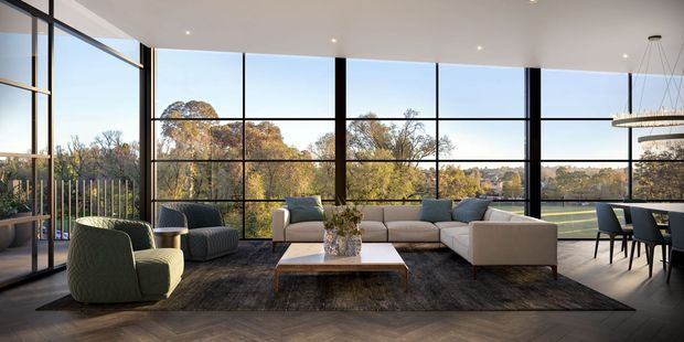 A sophisticated open-plan living room with floor-to-ceiling black-framed glazing overlooking mature trees. The room features a large neutral sectional sofa, sculptural lounge chairs, a low rectangular coffee table, dark area rug, dining table and chairs, recessed ceiling lights, and a contemporary linear pendant. Warm timber flooring and extensive glazing create a bright, refined interior.