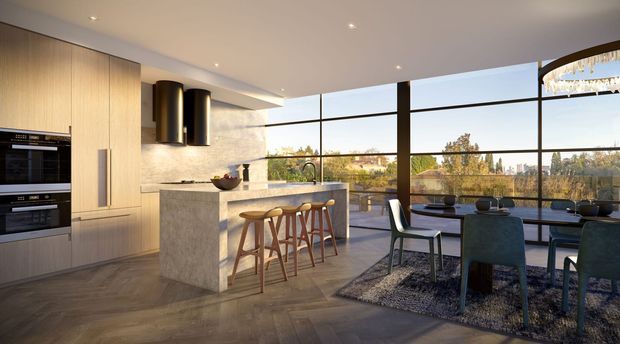 A refined open-plan kitchen and dining area with floor-to-ceiling black-framed glazing. The kitchen features pale timber cabinetry, integrated appliances, a stone island with timber bar stools, and dark cylindrical pendant lighting. A dining table with upholstered chairs sits on a textured rug beside the glazing, overlooking trees and neighbouring homes. The space is bright, warm, and contemporary with strong indoor-outdoor visual connection.