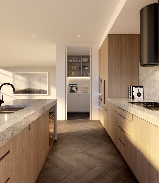 A warm, contemporary kitchen viewed down the main work aisle. The room features extensive light-to-mid wood cabinetry, pale stone benchtops, a large island with an undermount sink and dark curved tap, integrated appliances, open pantry shelving with concealed lighting, a textured pale splashback, and dark herringbone timber flooring. Recessed ceiling lighting and strong late-afternoon sunlight create a refined, inviting atmosphere.