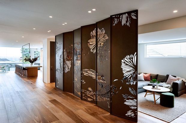 Open-plan contemporary interior featuring a large decorative dark metal or timber-toned screen partition with intricate botanical cut-out motifs. The partition separates a warm timber-floored dining and kitchen area from a bright living room with neutral furnishings, rug, coffee table and large windows. Recessed ceiling lighting and clean-lined detailing create a refined, modern atmosphere.