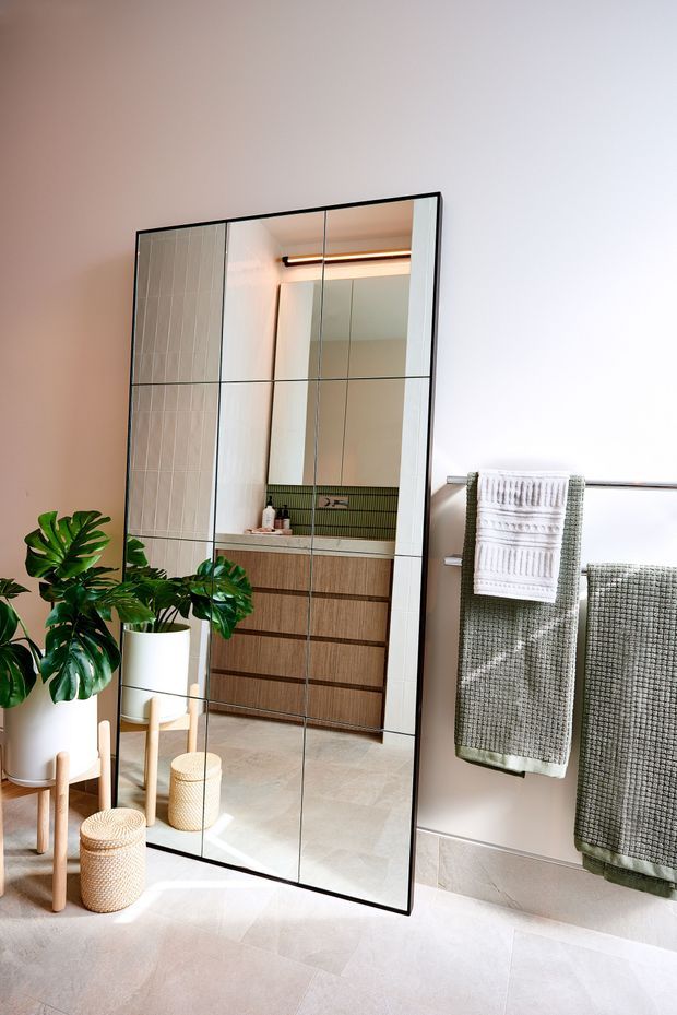 Bright contemporary bathroom featuring a large black-framed full-height mirrored panel, a floating timber vanity with integrated basin and green vertical-tile splashback visible in the reflection, pale stone-look floor and wall finishes, and a heated towel rail with folded towels. Large indoor plants and woven baskets add natural texture.