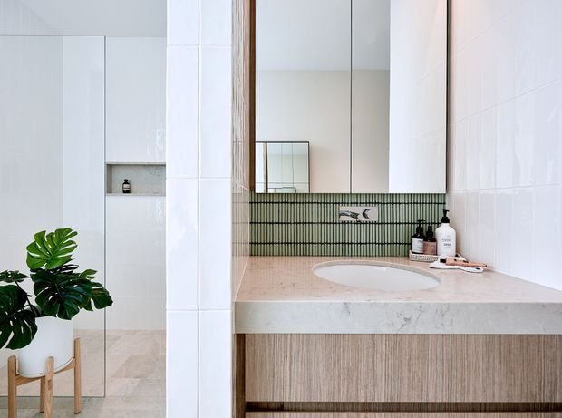 Bright contemporary bathroom featuring a pale stone vanity benchtop with an integrated oval basin, timber-look vanity cabinetry, white tiled walls, and a vertically ribbed green tile splashback. A large mirror, wall-mounted tapware, recessed shower shelving, and a potted plant complete the clean, refined space.