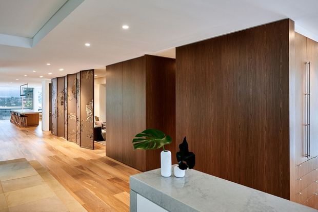 Open-plan contemporary interior with a warm timber floor and large dark timber wall panels forming a hallway and room dividers. A pale stone-topped island or counter occupies the foreground, with recessed ceiling lighting, built-in cabinetry, decorative screens, and glazing at the far end.