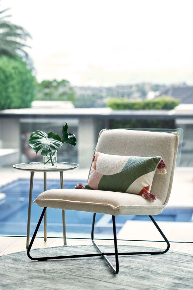 A bright, airy indoor seating vignette beside full-height glazing, featuring a cream upholstered lounge chair with slim black metal legs, a geometric multicoloured cushion, and a small round metal side table holding a glass vase with tropical leaves. A blue swimming pool and landscaped suburban setting are visible beyond the glazing.