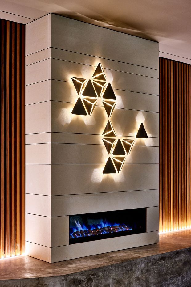 A dramatic contemporary interior feature wall with a built-in linear electric fireplace set into large-format grey stone or concrete-look panels. Vertical dark timber slat screens frame the wall on both sides, while geometric illuminated wall lights create a sculptural focal point above the fireplace. The space has warm ambient lighting, dark flooring, and a refined, moody atmosphere.