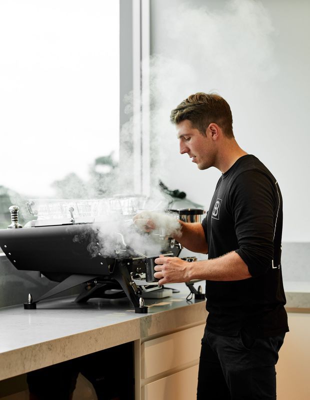 A commercial-style coffee preparation scene showing a barista operating a large black espresso machine on a pale stone countertop. Steam rises prominently from the machine, with light cabinetry below and bright glazing or a window in the background.