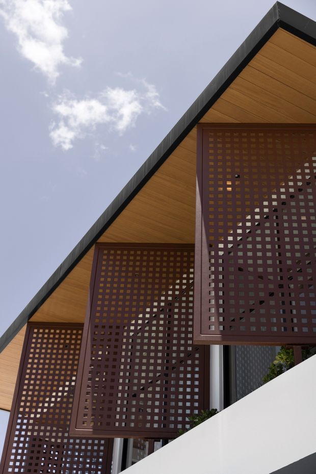 Upward-looking exterior architectural photograph of a contemporary multi-level facade. Repeating steep triangular roof or gable forms have warm timber soffits framed in dark metal, while projecting dark burgundy perforated lattice screens create patterned privacy panels across the glazing. A white lower wall and glimpses of plants appear at the base, against a pale blue sky.