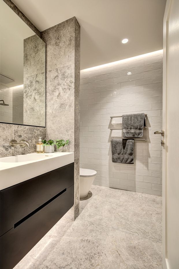 Contemporary bathroom with a floating dark vanity, white integrated basin, large illuminated mirror, pale marble-look wall and floor tiles, white brick-pattern wall tiles, and a wall-mounted towel rack. A toilet and partially screened shower area are visible.