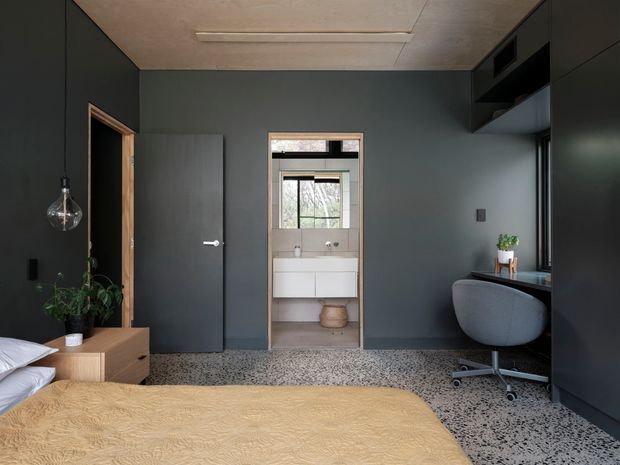 A compact, dark-toned bedroom with a low-profile bed, warm textured bedding, terrazzo flooring, built-in dark cabinetry, a small desk and swivel chair, and a pendant light. An open doorway reveals a bathroom with a wall-mounted vanity and window. The room has a restrained contemporary aesthetic with contrasting timber door trim and warm neutral accents.