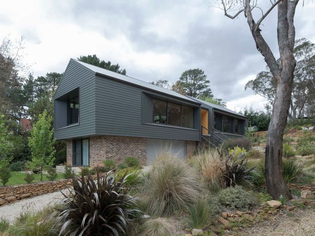 Exterior view of a contemporary split-level house set within a landscaped, rocky garden. The upper level is clad in dark horizontal weatherboards with large dark-framed windows, while the lower level combines exposed reddish brick with lighter grey wall surfaces. A small stair leads to the entry, and native grasses, spiky plants, stone edging, and mature trees surround the building.