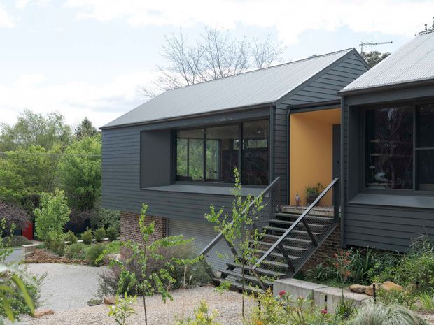 Exterior view of a contemporary multi-level home composed of dark horizontal cladding, a pitched metal roof, large dark-framed windows, and a projecting entry volume. A stairway leads through landscaped garden beds to the warmly lit entrance, with brickwork visible beneath the upper level.