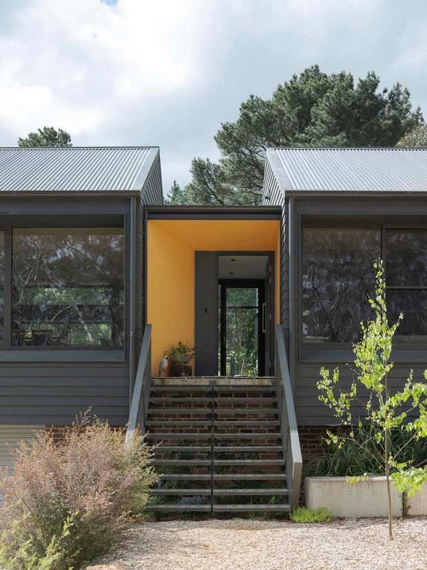 Front exterior view of a contemporary single-storey home composed of two dark horizontal-clad wings with steep corrugated metal gable roofs. A recessed entry porch with a vivid warm yellow-orange ceiling and wall sits between the wings, reached by a central flight of brick-and-metal stairs. Large dark-framed windows reflect surrounding trees, with sparse landscaping and gravel in the foreground.
