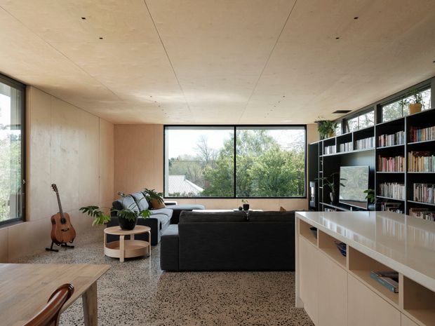 Warm, open-plan contemporary living room with a large dark sectional sofa facing a built-in black bookcase and media unit. Pale timber wall and ceiling panels, expansive black-framed windows, terrazzo flooring, indoor plants, and a guitar create a calm, natural connection to the leafy outdoors. A substantial pale built-in joinery unit occupies the foreground.