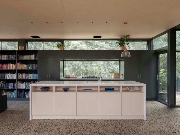 Contemporary kitchen with a large pale island featuring integrated open shelving and cabinetry, dark full-height cabinetry, a broad window splashback, and extensive glazing looking onto leafy surroundings. A built-in bookshelf occupies the left side, while terrazzo-style flooring and restrained pendant lighting complete the refined interior.