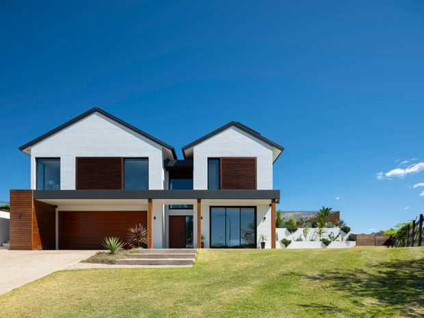 Front exterior view of a contemporary two-storey residential house with two prominent pitched gable roof forms, white rendered walls, dark trim, timber-toned privacy screens, large glazed openings, and an integrated garage. A broad lawn, driveway, front steps, and landscaped planting are visible in the foreground.