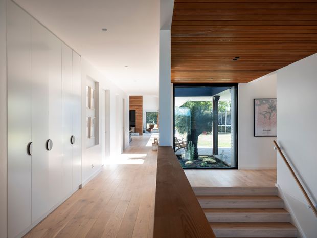 Bright contemporary residential hallway and stairway with extensive white built-in cabinetry, pale timber flooring, a dark timber stair balustrade, and a warm slatted timber ceiling. Large glazing at the stair landing connects visually to a landscaped outdoor area, while framed artwork and recessed lighting add understated detail.