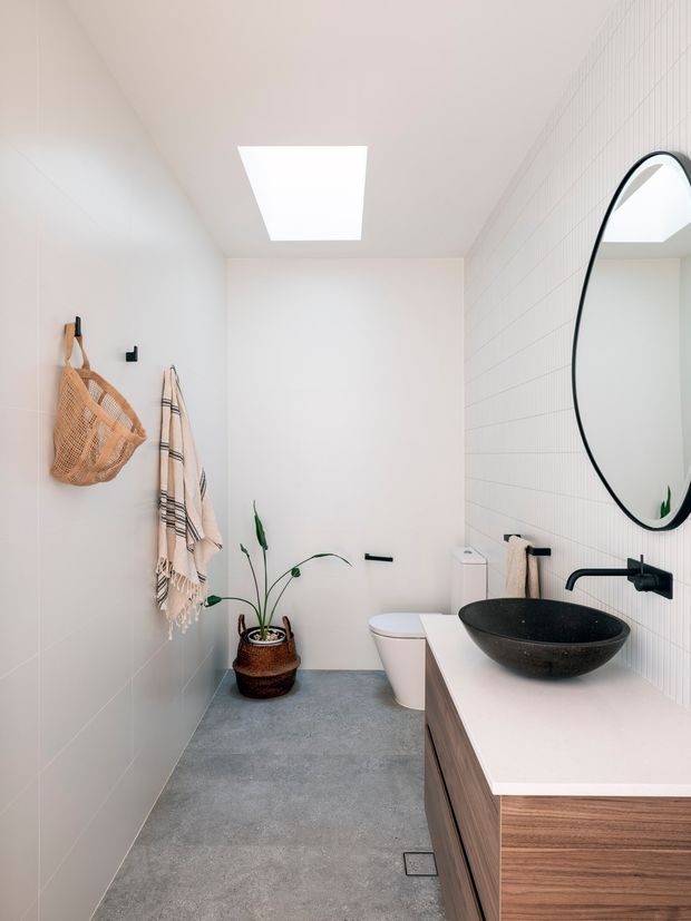 Bright, narrow contemporary bathroom with white tiled walls, a large rounded black-framed mirror, black vessel basin and wall-mounted tap on a white benchtop, and a timber vanity with integrated storage. A toilet, skylight, grey floor tiles, hanging towels, woven basket and potted plant complete the calm, minimalist fit-out.