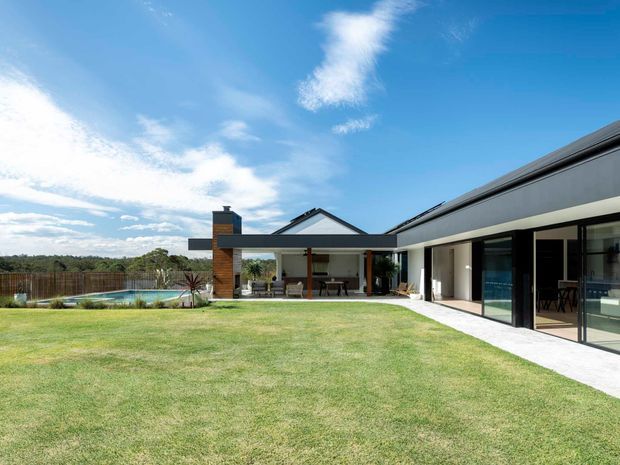A single-storey contemporary residence viewed from a broad lawn, with a long dark-clad wing, extensive floor-to-ceiling glazing, a covered outdoor entertaining area, and a pitched-roof section around a brick chimney. A swimming pool and landscaped boundary are visible to the side.