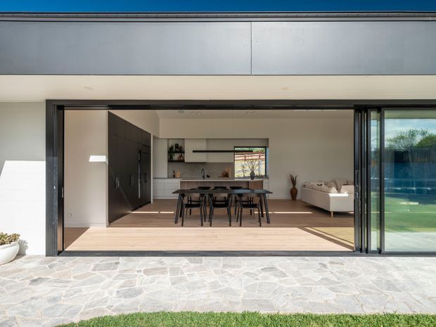 View from an outdoor paved area through fully opened sliding glass doors into a bright open-plan interior. The space combines a contemporary kitchen with dark fixed cabinetry and island, a dining area with a timber table and chairs, and a living area with a light sofa. Light timber flooring, pale walls, extensive black-framed glazing, and a spacious indoor-outdoor connection are prominent.
