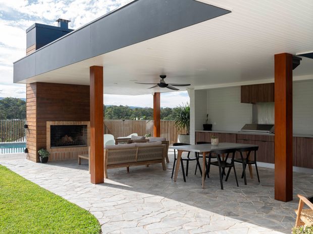 A contemporary covered outdoor entertaining area adjoining a modern home, with timber posts, a broad flat roof, stone-paved flooring, built-in timber-clad fireplace, outdoor lounge seating, dining table, and an integrated outdoor kitchen. The space overlooks a landscaped yard and distant greenery, with a swimming pool partially visible at the side.