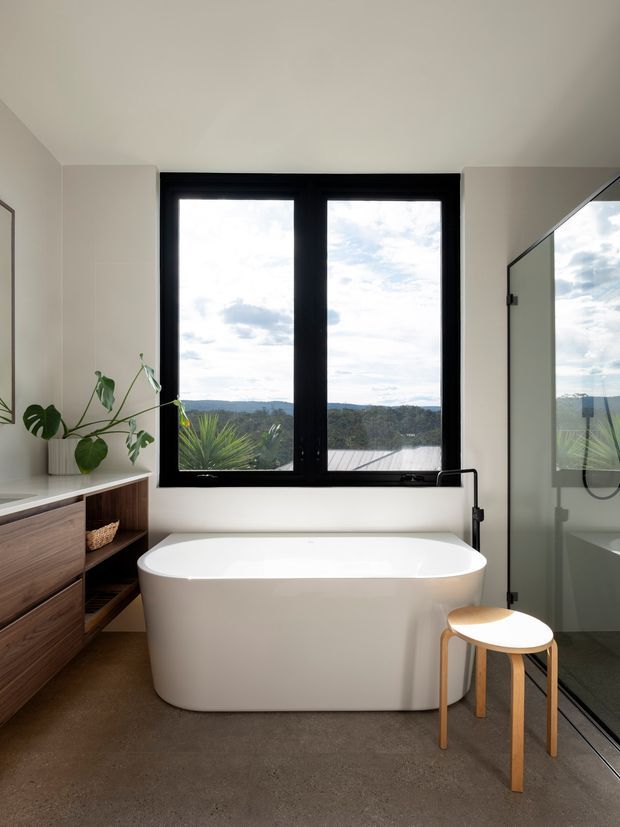 Contemporary bathroom with a freestanding white bathtub positioned beneath a large black-framed window overlooking a wooded landscape. A glass shower enclosure, wall-mounted bath tap, timber vanity cabinetry, indoor greenery, and a small timber stool complete the calm, minimalist space.