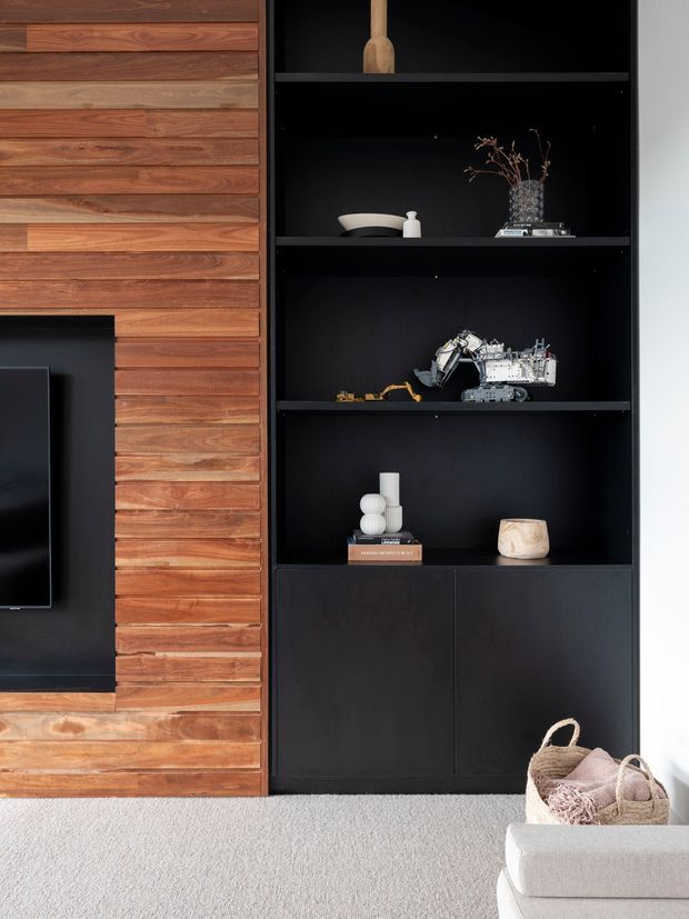 Close-up interior view of a contemporary living room media wall, featuring warm horizontal timber wall cladding, a black recessed television surround, and full-height matte-black built-in shelving and cabinetry displaying decorative objects. A light neutral sofa and textured carpet are partially visible in the foreground.