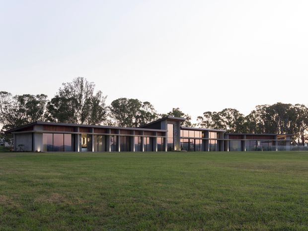 A wide exterior view of a long, low-slung contemporary building set behind an expansive lawn. The building has a strongly horizontal composition, dark slim roof planes, extensive floor-to-ceiling glazing, and a taller central vertical volume. Mature trees form a backdrop, and the image is photographed in soft evening light.