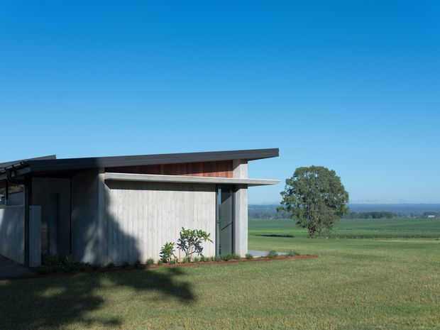 A low, modern rural building viewed from outside, with a broad asymmetrical flat or very shallow-pitched roof, exposed concrete walls, dark framed glazing, and warm timber cladding beneath the roofline. The structure sits in an open mown grass setting with farmland and a distant tree line beyond, under a clear blue sky.