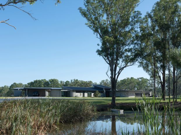 Wide exterior view of a low, elongated contemporary building set within a spacious landscaped rural setting. The dark-clad structure has a mostly flat or very low-pitched roof, broad glazing, and covered outdoor areas. Mature eucalyptus trees, lawn, native grasses, and a reflective pond dominate the foreground and reinforce the natural Australian landscape context.