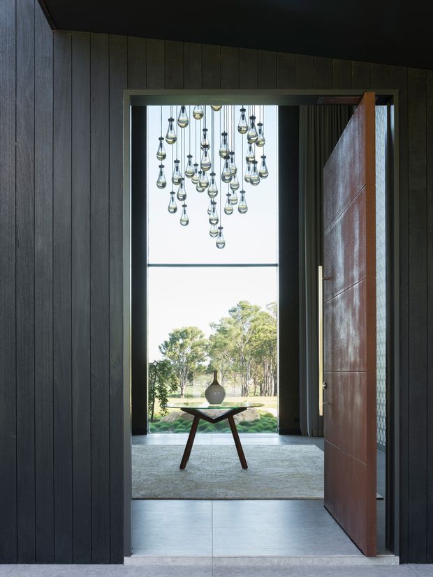 A dramatic residential entryway viewed from a dark timber-lined hall. A large pivot door is open toward a bright glazed entrance, with a sculptural hanging cluster of pendant lights above a small console table and vase. The space opens onto a landscaped rural-looking exterior, while the interior uses dark vertical wall panelling, large pale floor tiles, and a warm timber door.