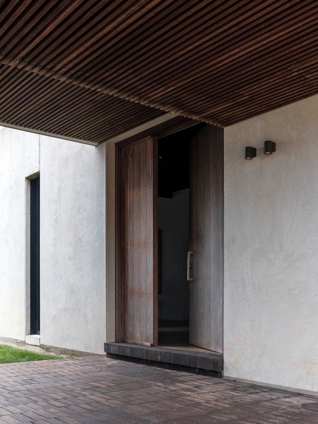 Close exterior view of a contemporary entry facade with a tall dark timber pivot door set into pale textured plaster walls. A deep overhead canopy is lined with closely spaced dark timber battens, complemented by minimalist black wall sconces. The entrance opens onto a dark brick-paved forecourt, with a narrow vertical window beside the door and a small strip of lawn at the edge.