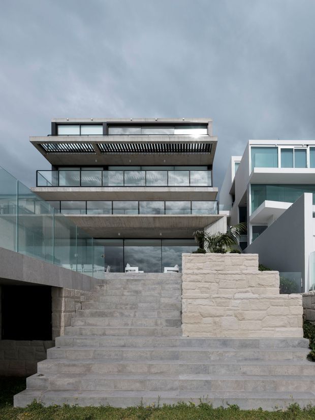 Exterior view of a multi-level contemporary residential building with broad cantilevered balconies, extensive floor-to-ceiling glazing, glass balustrades, a louvred upper balcony, and wide pale stone or concrete steps leading up to the house. The facade is set against a dramatic overcast sky, with a neighbouring modern residence visible on the right.