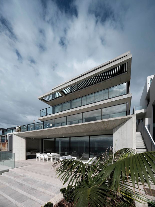 Exterior photograph of a contemporary multi-storey concrete residence with extensive floor-to-ceiling glazing, projecting terraces, glass balustrades, and a prominent slatted roof overhang. Broad pale stone or concrete steps lead to a large outdoor terrace with dining furniture, while tropical palms occupy the foreground. The building sits on a stepped or sloping site beneath a dramatic cloudy sky.