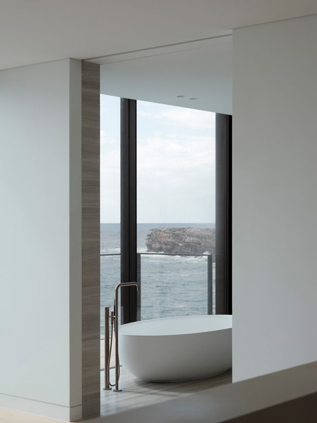 Minimalist bathroom with a freestanding white bathtub positioned beside floor-to-ceiling glazing, overlooking a rugged ocean coastline. The room has smooth white walls, dark slender window frames, a warm grey-brown wood-look feature surround, and a pared-back contemporary fit-out.