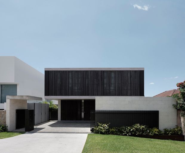 Street-facing view of a contemporary two-storey residence with a stark rectangular facade. The upper level is clad in dark vertical slatted panels, contrasted with pale rendered and masonry walls below. A recessed dark entry and garage gate sit behind a paved driveway, with a low black slatted boundary fence, clipped planting, and lawn in the foreground. Clear blue sky and neighbouring homes frame the property.