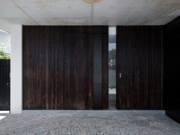 A dark, minimalist architectural entrance beneath a broad raw concrete canopy. The facade is dominated by tall vertical dark timber panels and a matching pivot door, framed by pale concrete columns. A narrow opening reveals the street beyond, while the foreground is paved with small grey cobblestones. The composition emphasizes strong horizontal and vertical lines, tactile materials, and a restrained contemporary character.