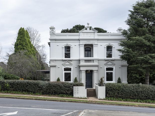 Street-facing view of a restored two-storey heritage residence with a symmetrical pale grey rendered facade, ornate classical detailing, decorative window surrounds, balustrades, parapet elements, and a central entrance. Mature trees and hedges frame the property, with a road in the foreground.