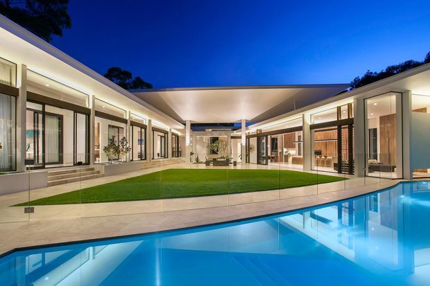 Twilight exterior view of a low-slung contemporary residence arranged around a landscaped courtyard and bright swimming pool. The home features extensive floor-to-ceiling glazing, clean horizontal rooflines, pale rendered walls, warm interior lighting, and a frameless glass pool barrier.