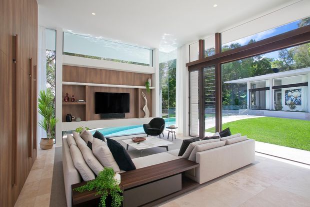 Bright contemporary living room with floor-to-ceiling glazing and large timber-framed sliding doors opening toward a lawn and pool. The room features a built-in timber media wall, low-profile cream sectional seating, dark timber coffee table, black accent chair, pale stone or tile flooring, and a clean white ceiling. A modern pavilion-like structure is visible outside.